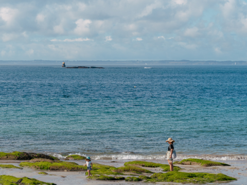 Una madre y su hijo pasean por la playa rocosa cerca de Quiberon, Bretaña, Francia, con el mar y un faro al fondo.