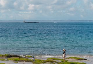Una madre y su hijo pasean por la playa rocosa cerca de Quiberon, Bretaña, Francia, con el mar y un faro al fondo.