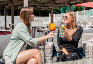 Dos mujeres disfrutan bebidas coloridas y sol en una terraza en Flower Camping Le Bois d’Amour, Bretaña, Francia.