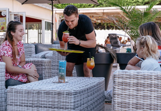 Un camarero sirve coloridas bebidas a huéspedes sonrientes en la terraza de Flower Camping Le Bois d'Amour.
