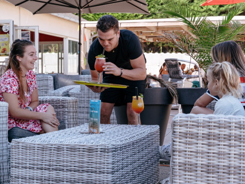 Een kelner serveert kleurrijke drankjes aan lachende gasten op het terras van Flower Camping Le Bois d'Amour.