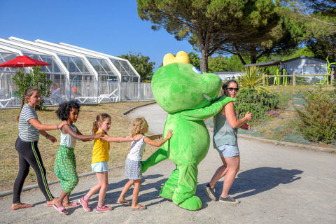 Des enfants et une adulte font une farandole derrière une mascotte verte au Flower Camping Le Bois d'Amour en Bretagne.