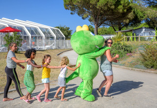 Kinder folgen einer Erwachsenen und einem grünen Maskottchen im Flower Camping Le Bois d'Amour, Bretagne, Frankreich.