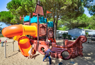 Children play on a colorful pirate ship playground with slide, set on sand at a holiday park surrounded by trees.