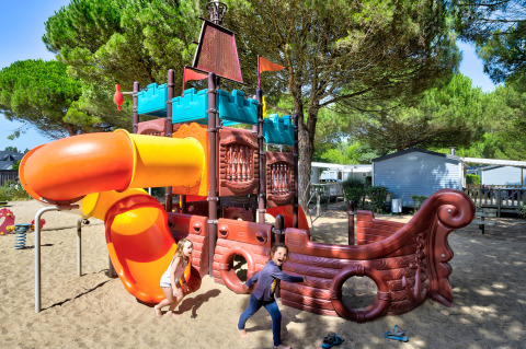Children play on a colorful pirate ship playground with slide, set on sand at a holiday park surrounded by trees.