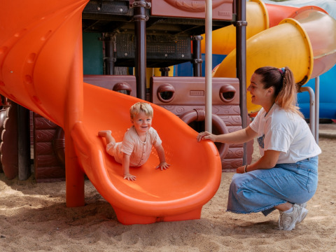 Un bambino scende da uno scivolo arancione mentre un adulto sorride al Flower Camping Le Bois d'Amour.