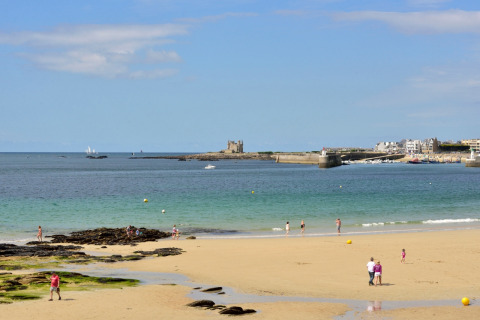 People are enjoying a sunny beach day near Quiberon in Brittany, France, with a castle and boats in view.