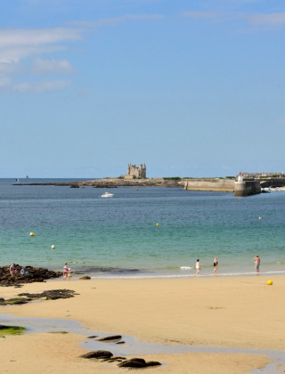 Folk nyder en solrig dag på stranden i nærheden af Quiberon med udsigt over havet og en borg i baggrunden.