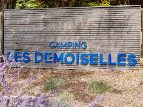 Indgangsskilt til Camping Les Demoiselles, feriepark i Pays de la Loire, Frankrig, omgivet af natur.