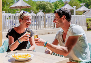 Twee blije mensen proosten met drankjes en eten frietjes op Camping Demoiselles Plage in Pays de la Loire.