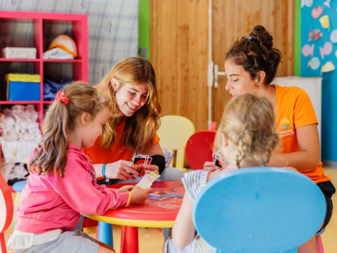 Two children and two adults play cards at a table in the indoor playroom at Camping Demoiselles Plage.