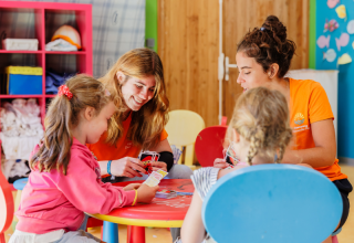 Two children and two adults play cards at a table in the indoor playroom at Camping Demoiselles Plage.