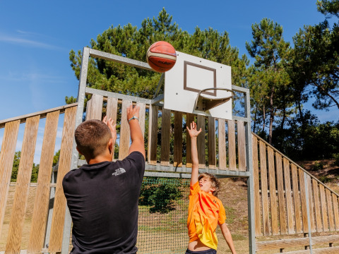 Twee mensen spelen buiten basketbal aan Camping Demoiselles Plage, Pays de la Loire, Frankrijk, onder blauwe hemel.