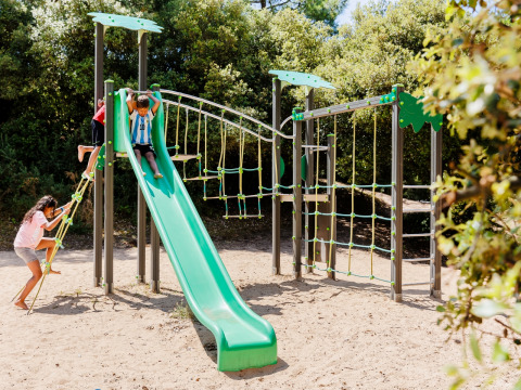 Children playing on a green slide and climbing frame in a sandy playground surrounded by trees in France.