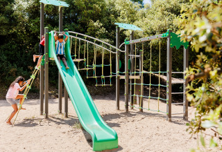 Children playing on a green slide and climbing frame in a sandy playground surrounded by trees in France.
