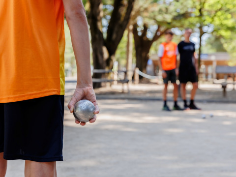 Person in orange shirt holding a pétanque ball, ready to play on an outdoor court in Pays de la Loire, France.