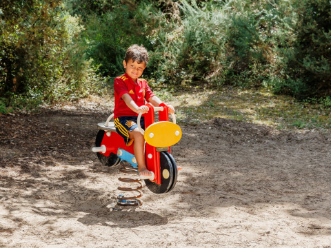 Bambino con maglia rossa su dondolo colorato in un parco giochi all’aperto a Camping Demoiselles Plage, Francia.