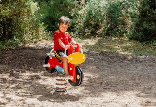 Bambino con maglia rossa su dondolo colorato in un parco giochi all’aperto a Camping Demoiselles Plage, Francia.