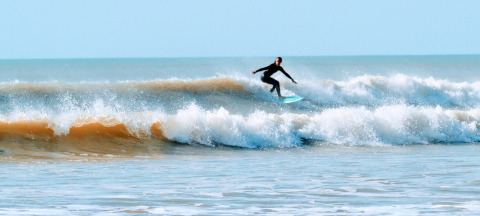A surfer rides a wave near Saint-Hilaire-de-Riez, Pays de la Loire, France, with clear blue skies overhead.