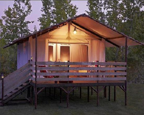 Tenda safari illuminata presso Cabane Lodge con veranda in legno e rampa, circondata da alberi al tramonto.