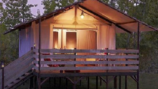 Tienda safari iluminada en Cabane Lodge con terraza de madera y rampa, rodeada de árboles al anochecer.