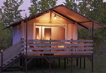 Tienda safari iluminada en Cabane Lodge con terraza de madera y rampa, rodeada de árboles al anochecer.