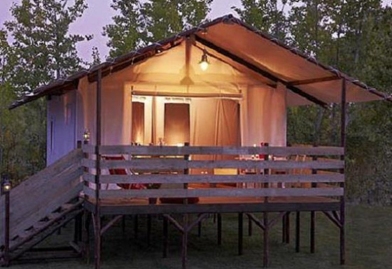 Tenda safari illuminata presso Cabane Lodge con veranda in legno e rampa, circondata da alberi al tramonto.