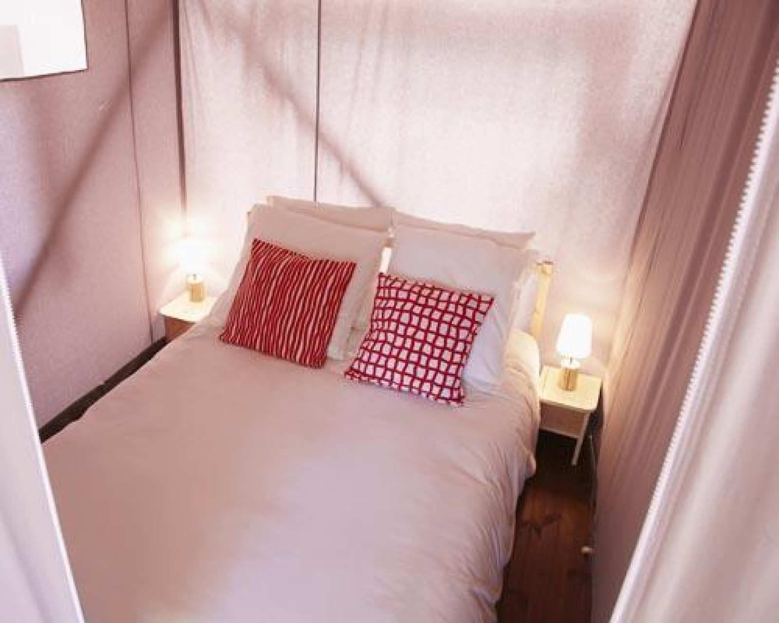Foto van een knus bed met rode kussens en nachtlampjes in een safari tent met stoffen wanden.