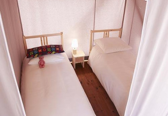 Interior de una tienda safari con dos camas individuales, mesita de noche con lámpara y suelo de madera.