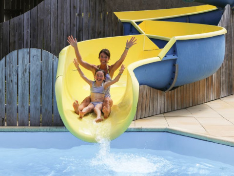 Una mujer y una niña en bañadores bajan sonrientes por el tobogán amarillo en Flower Camping Les Dunes.