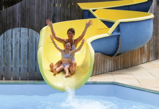 Una mujer y una niña en bañadores bajan sonrientes por el tobogán amarillo en Flower Camping Les Dunes.