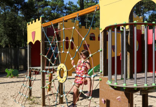 A child climbing on a rope net at a castle-themed playground with sand at Flower Camping Les Dunes, France.