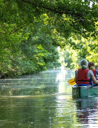 To personer padler i en kano på en stille flod omgivet af frodig grøn skov nær Longeville-sur-Mer.