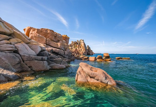 Felsige Küste und klares Wasser am Flower Camping Port de la Chaîne in der Bretagne, Frankreich, unter blauem Himmel.