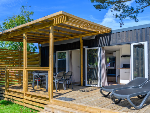 Glampingaccommodatie met houten terras, ligstoelen en overdekte eethoek bij zonnig weer en groene bomen.