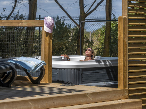 Vrouw ontspant in een buitenjacuzzi bij een glamping, omringd door houten terras en zonnige natuur.