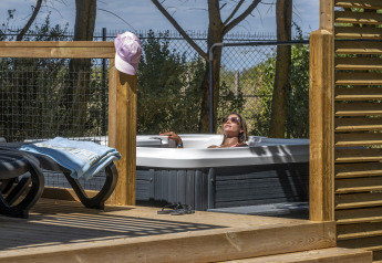 Kvinde slapper af i et udendørs spabad ved glamping, omgivet af træterrasse og solrige omgivelser.