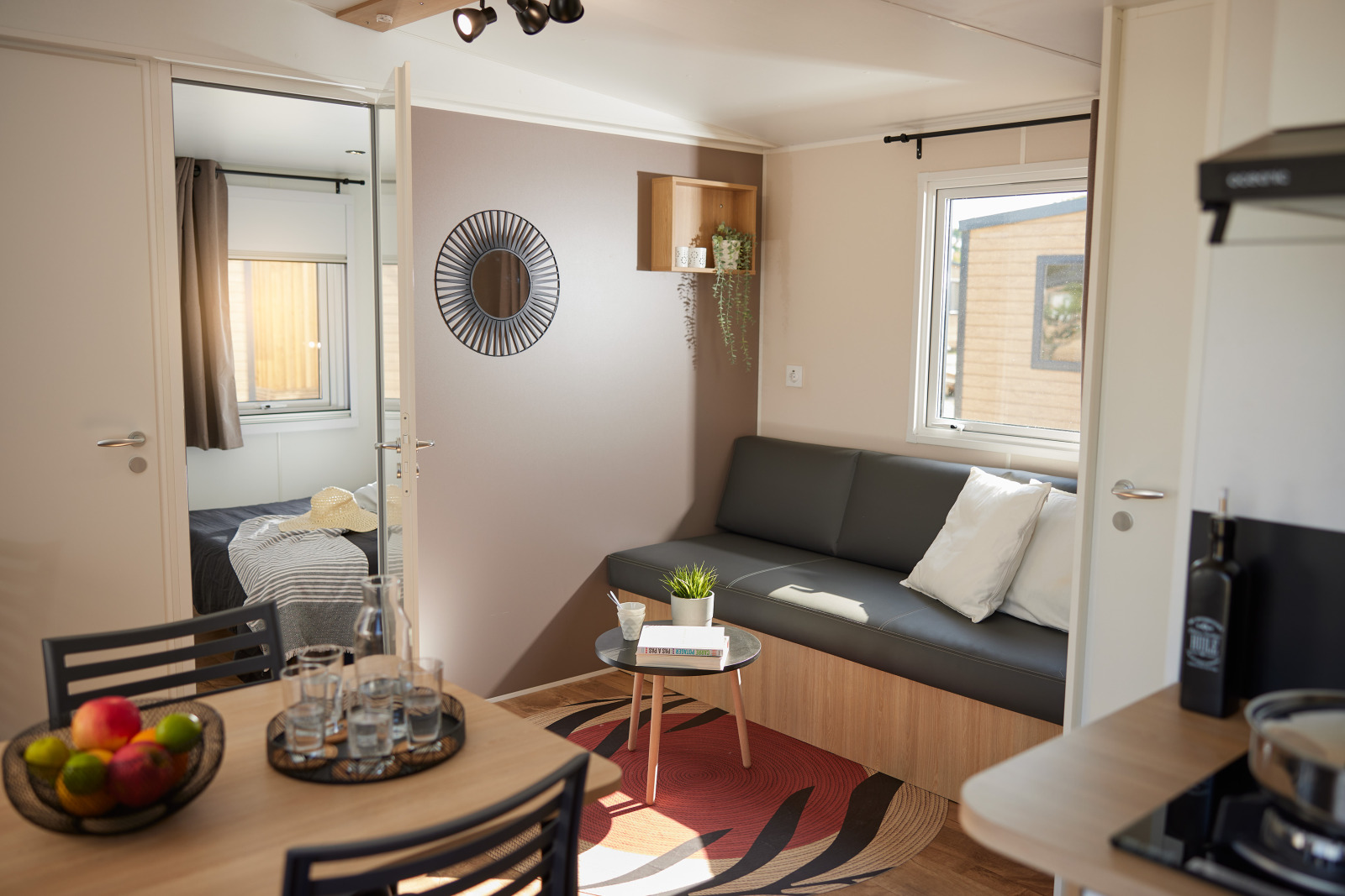 Moderne glampingaccommodatie met woonkamer, grijze bank, eettafel en zicht op de slaapkamer.