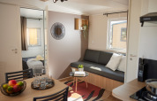 Moderne glampingaccommodatie met leefruimte, grijze zetel, eettafel en zicht op de slaapkamer.