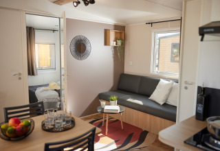 Moderne glampingaccommodatie met leefruimte, grijze zetel, eettafel en zicht op de slaapkamer.
