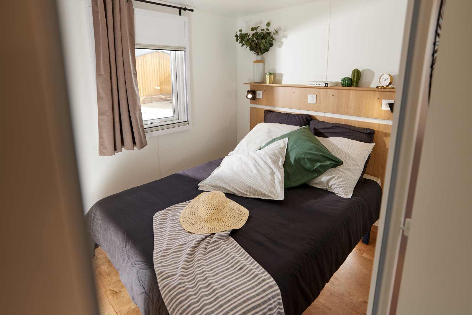Sfeervolle glamping slaapkamer met tweepersoonsbed, groen kussen, strohoed en natuurlijk daglicht.