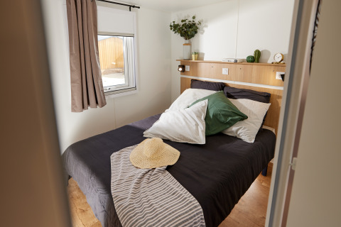 Gezellige glamping slaapkamer met een tweepersoonsbed, groene kussen, strohoed en natuurlijk licht.