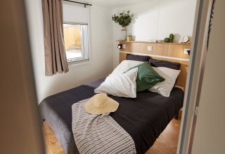 Gemütliches Glamping-Schlafzimmer mit Doppelbett, grünes Kissen, Strohhut und viel Tageslicht.