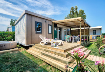 Et moderne Homeflower PREMIUM Jacuzzi-lodge med træterrasse, solsenge og udendørs boblebad, omgivet af grønne planter.