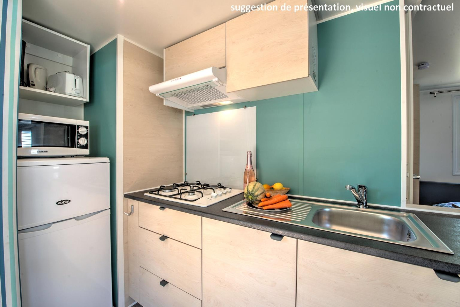 Compacte glampingkeuken met gasfornuis, spoelbak, koelkast, magnetron en een bord verse groenten.