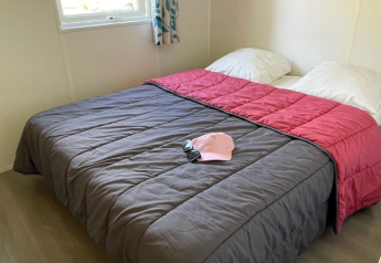 Habitación con cama doble en Mobitoile Standaard de Flower Camping Les Ondines, Francia, con gafas de sol y gorra rosa en la cama.