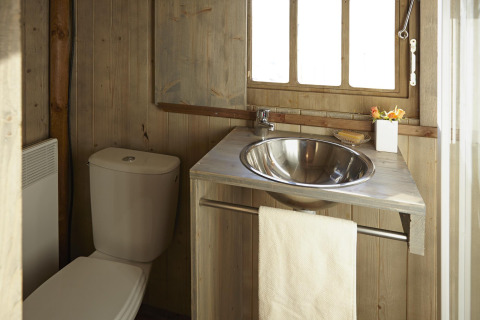 Piccolo bagno in alloggio glamping con pareti in legno, lavandino in acciaio e toilette vicino alla finestra.