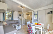 Moderne glamping-accommodatie met lichte keuken, eettafel, grijze bank en ramen met gordijnen.