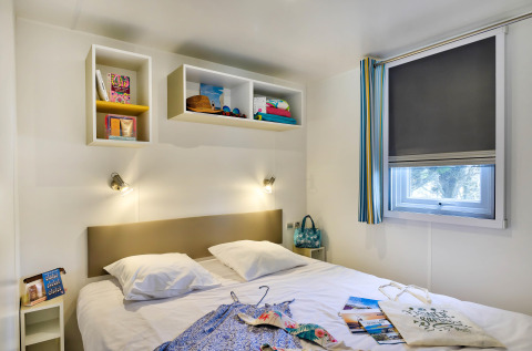 Moderne glamping slaapkamer met dubbel bed, wandrekjes, raam met rolgordijn en kleurrijke accessoires.