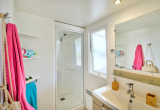 Salle de bain lumineuse avec douche, fenêtre, étagères et serviettes colorées dans un hébergement glamping.